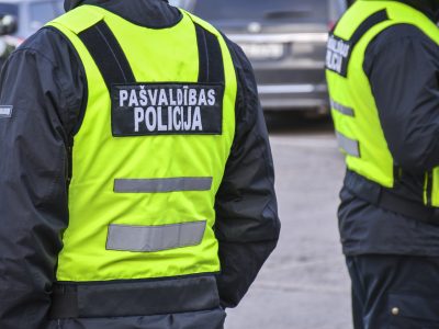 Divi pašvaldības policisti redzami no muguras, tērpušies melnās jakās un spilgti dzeltenās atstarojošās vestēs. Uz kreisā policista vestes ir uzraksts "PAŠVALDĪBAS POLICIJA".