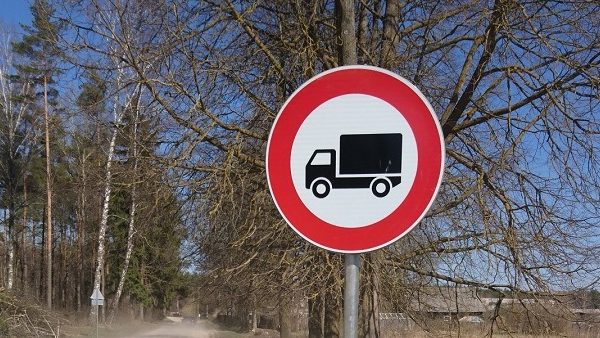 Apaļa ceļa zīme ar sarkanu apmali un melnu kravas automašīnas siluetu baltā fonā, kas norāda kravas automašīnu satiksmes aizliegumu. Fonā kaili koki, zilas debesis un grants ceļš.