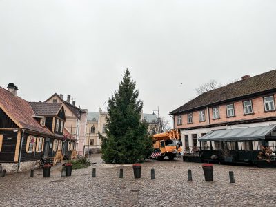 Liela Ziemassvētku egle Kuldīgas Rātslaukumā, tai blakus oranža celtņa mašīna, ko ieskauj vēsturiskas ēkas. Uz vienas no tām redzams uzraksts "RESTORĀNS".