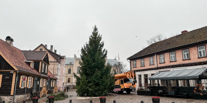 Liela Ziemassvētku egle Kuldīgas Rātslaukumā, tai blakus oranža celtņa mašīna, ko ieskauj vēsturiskas ēkas. Uz vienas no tām redzams uzraksts "RESTORĀNS".