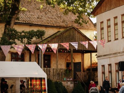 Festīva aina Alsungā ar senām ēkām un cilvēkiem, virs kuras karājas pleda raksta karodziņu virtene. Priekšplānā balta telts, bet pie koka nojumes redzama zīme ar uzrakstu "i ieeja pils".