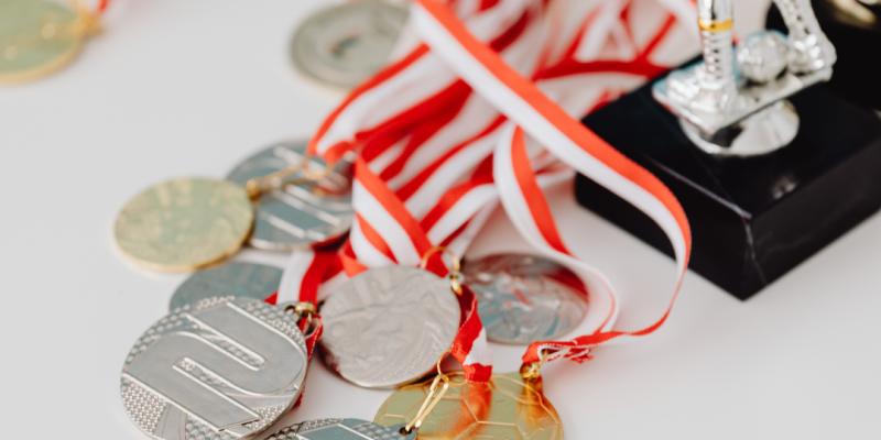 Uz balta fona redzamas sudraba un zelta medaļas ar sarkanbaltām lentēm