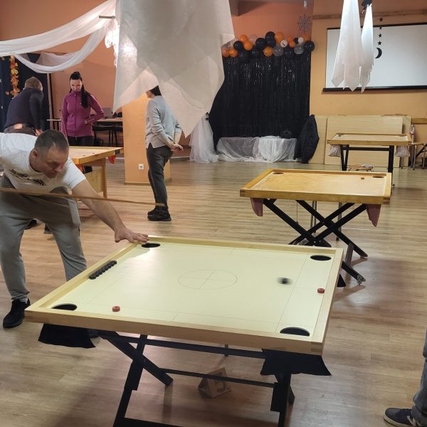 Vīrietis baltā T-kreklā spēlē novusu, tēmējot ar kiju pa melnu ripu uz gaiša galda. Viņam blakus labajā pusē stāv vecāks vīrietis zilā jakā ar rūtainu kreklu, turot kiju un vērojot spēli.