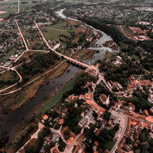 Gaisa skats uz Kuldīgas pilsētu ar Ventas upi, veco ķieģeļu tiltu un plašo Ventas rumbu. Abpus upei redzamas ēkas ar sarkanīgiem jumtiem, ko ieskauj zaļi lauki un koki.