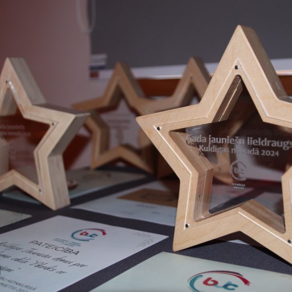 Vairāki koka zvaigznes formas apbalvojumi ar caurspīdīgiem centriem un sertifikāti uz galda. Uz priekšējā apbalvojuma ir uzraksts: "Gada jauniešu lieldraugs Kuldīgas novadā 2024".