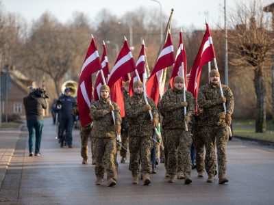 Vairāki vīrieši militārās maskēšanās formās soļo pa ielu, nesot paceltus Latvijas karogus. Fonā redzams orķestris