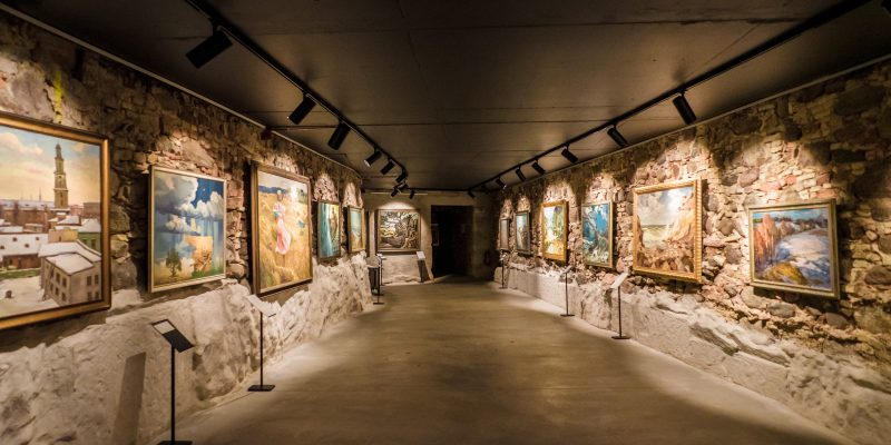 Plaša gleznu izstāde šaurā, garenā telpā ar neapstrādātām akmens sienām un tumšu griestiem, kur gleznas izgaismo prožektori.