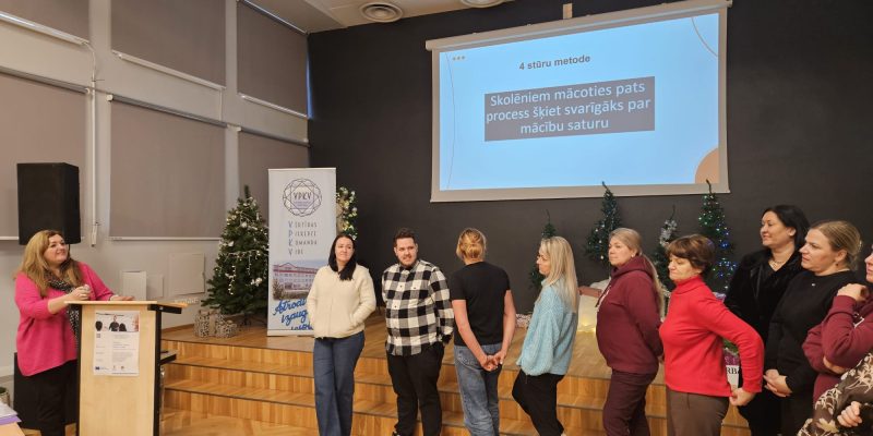 Sieviete rozā džemperī runā no pults auditorijas priekšā, kurā stāv vairākas sievietes un vīrieši.