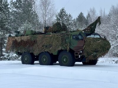 Bruņots zaļš militārais transportlīdzeklis ar maskēšanās tīklu stāv uz sniega klātas zemes ziemas mežā.