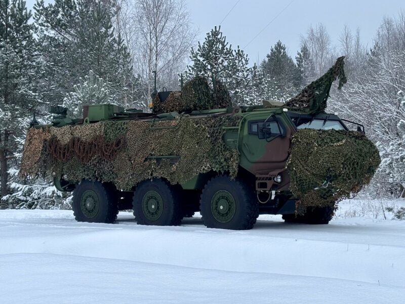 Bruņots zaļš militārais transportlīdzeklis ar maskēšanās tīklu stāv uz sniega klātas zemes ziemas mežā.