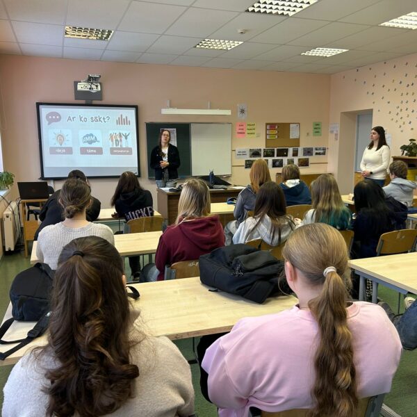 Klasē pie prožektora ekrāna, kurā redzams uzraksts "Ar ko sākt?", "Ideja", "Tēma", stāv sieviete, prezentējot studentiem, kas sēž pie galdiem. Otra sieviete stāv klases labajā pusē.