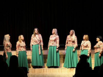 Septiņas sievietes saskaņotos tērpos – ziedu raksta blūzēs un garos, zaļos svārkos – uzstājas uz skatuves. Viņas stāv rindā pret tumšu fonu, dažas skatās viena uz otru, dažas uz priekšu, un viena sieviete dzied ar atvērtu muti. Priekšplānā redzami skatītāju silueti.