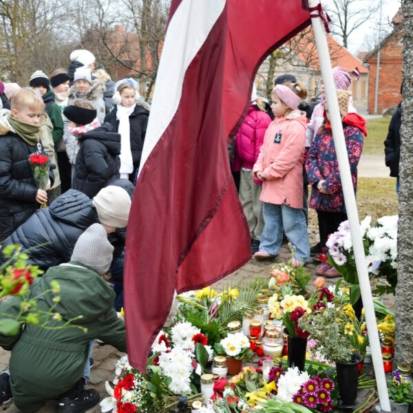 Bērni noliek ziedus un iedegtas sveces pie akmens pieminekļa, kura pakājē jau izvietoti daudzi ziedi un svecītes. Blakus plīvo Latvijas karogs. Fonā stāv vēl citi bērni un pieaugušie. Komunistiskā genocīda upuru piemiņas pasākumi novadā