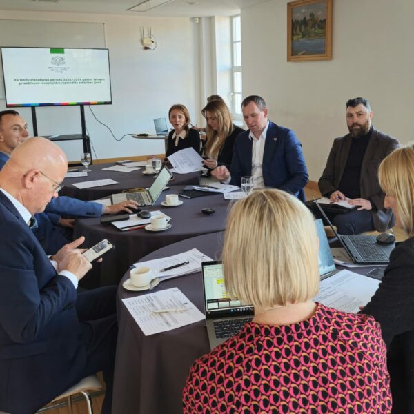 Sanāksme ar aptuveni desmit cilvēkiem, kas sēž pie liela galda, daži strādā ar klēpjdatoriem un pārskata dokumentus. Liels ekrāns rāda tekstu "ES fondu plānošanas periods 2028-2034 gadam Latvijā: priekšlikumi investīcijām reģionālās attīstības jomā".