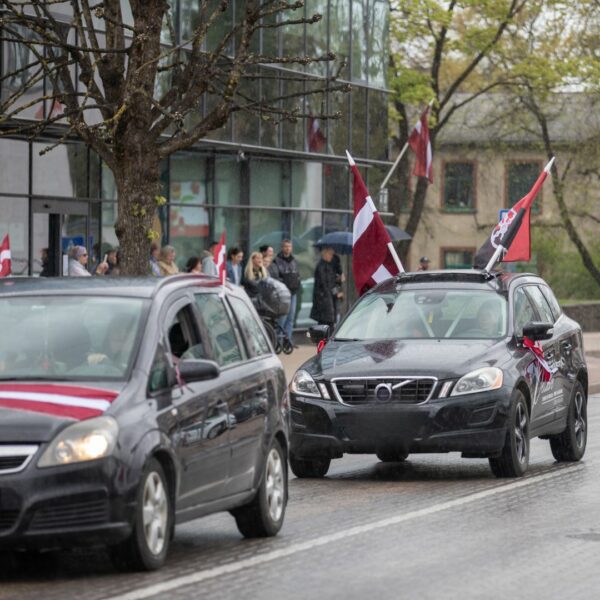 Mitrā ielā brauc divas melnas automašīnas, rotātas ar Latvijas karogiem. Priekšējais melnais apvidus auto uz jumta nes divus Latvijas karogus un sarkanmelnu karogu ar simbolu, uz tā sāniem ir uzraksts "Paldies visiem". Aizmugurējā automašīna ir daļēji redzama ar sarkanbaltsarkanu lentu. Fonā, uz ietves, stāv cilvēku pūlis, daži zem lietussargiem, vērojot notiekošo lietainā laikā.
