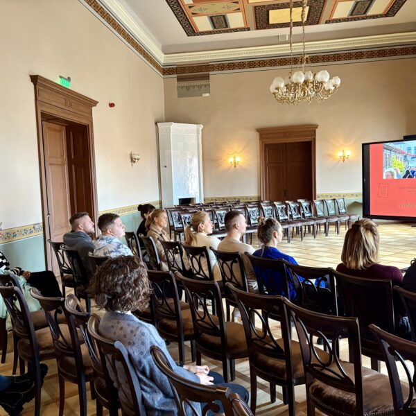 Grupa cilvēku sēž greznā zālē un skatās uz lielu ekrānu, kurā redzams ielas attēls