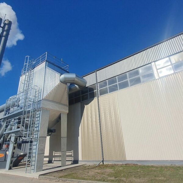 Industriāla ēka ar diviem augstiem pelēkiem skursteņiem, no viena nāk dūmi. Pie ēkas redzama ventilācijas sistēma un pelēks atkritumu konteiners ar oranžu uzrakstu "ALKO" gaiši zilās debesīs ar vienu mākoni.