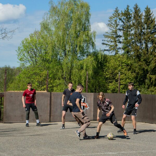 Pieci jauni vīrieši spēlē ielu futbolu uz āra asfalta laukuma ar koka sienām. Ēdolē notiks kausa izcīņa ielu futbolā