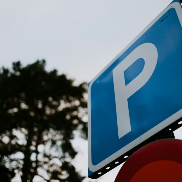 Zila taisnstūra stāvvietas zīme ar baltu burtu "P", redzama no apakšas pret pelēkām debesīm, ar izplūdušu tumšu koku fonā un daļēji redzamu sarkanu zīmi zem tās.