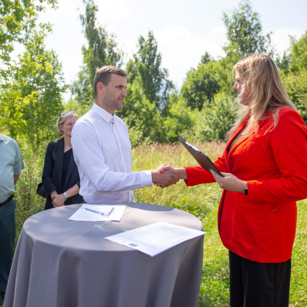 Vīrietis baltā kreklā un sieviete sarkanā žaketē - Kuldīgas novada domes priekšsēdētāja - paspiež rokas brīvā dabā, stāvot pie apaļa galda ar dokumentiem, tostarp vienu ar Eiropas Savienības simbolu.