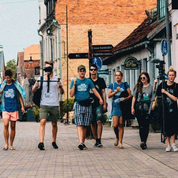 Seši jauni cilvēki iet pa bruģētu pilsētas ielu. Daži valkā T-kreklus ar uzrakstu "MEDIA SHARKS", un viens valkā T-kreklu ar uzrakstu "BAUNPILS". Divi no viņiem nes mediju aprīkojumu: viens tur kameras stabilizatoru, otrs – lielu statīvu ar mikrofonu. Fona ēkām ir vecas fasādes, un uz vienas ir uzraksts "SALONFRIZĒTAVA RITA anno 1964". Ielu zīme norāda uz "Annas baznīca 0.3", "WC 0.1", "Viegli uzliekas mašīna" un "BAUNPILS".