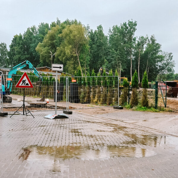 Lietainā dienā slapja bruģēta teritorija ar peļķēm. Aiz pagaidu metāla žoga strādnieki dzeltenās vestēs pie zila mini-ekskavatora ar uzrakstu "RŪTA" un asfalta kaudzēm. Blakus stāv ceļa zīme "Darbi uz ceļa" (sarkans trīsstūris ar strādnieka simbolu) un balta bultas zīme, kas norāda pa labi. Fonā redzamas koku audzes, industriālas ēkas un lieli būvgružu konteineri.