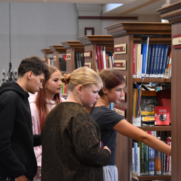Četri jaunieši, divas meitenes un divi zēni, aplūko grāmatas koka grāmatu plauktos bibliotēkā. Meitene labajā pusē sniedzas pēc grāmatas, kas atrodas zemākā plauktā. Uz viena no plauktiem ir uzraksts "Literatūrmīlne".