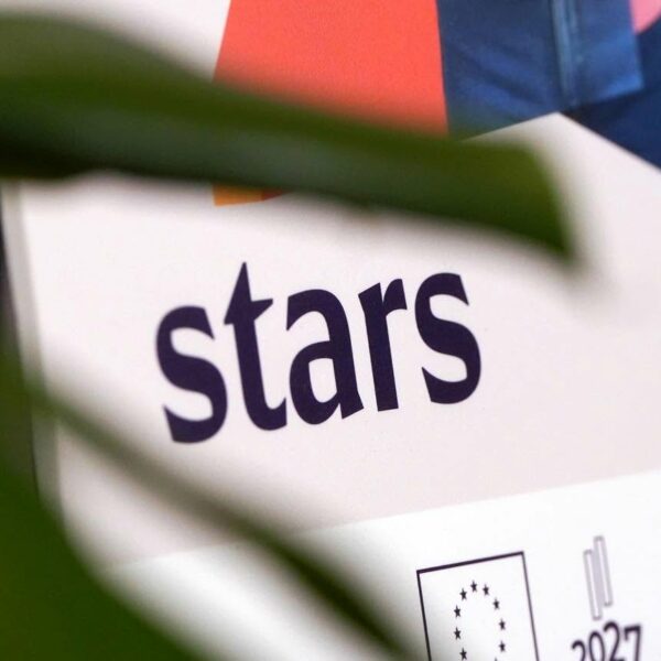 Balts plakāts vai stends ar vārdu "stars" tumši violetā krāsā. Zem tā redzams ES karoga logo, skaitlis "2027" un divi ģerbonim līdzīgi logo. Daļēji aizsegts ar zaļu augu lapām.