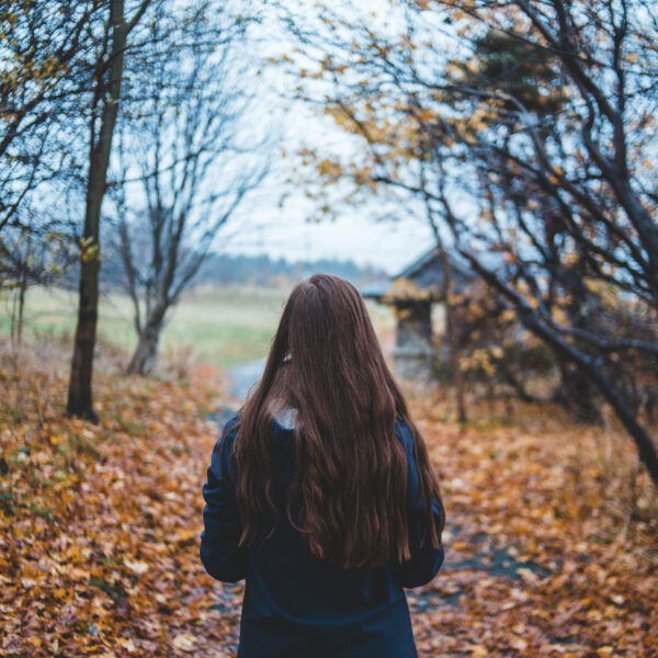 Persona no mugurpuses ar gariem brūniem matiem iet pa rudens taku, kas noklāta ar kritušām oranžbrūnām lapām, ar kailiem kokiem un lauku koka būdiņu fonā.